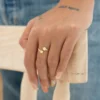 Una mano apoyada sobre una silla de madera, lleva un anillo dorado en forma de estrella y una pulsera verde con cuentas. La mano tiene un tatuaje que dice "SELF MADE". La persona lleva puesto un pantalón de mezclilla azul, resaltando la elegancia de Joyas en Oro Laminado 18K.