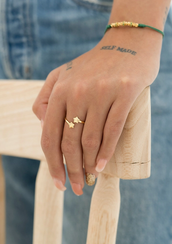 Una mano apoyada sobre una silla de madera, lleva un anillo dorado en forma de estrella y una pulsera verde con cuentas. La mano tiene un tatuaje que dice "SELF MADE". La persona lleva puesto un pantalón de mezclilla azul, resaltando la elegancia de Joyas en Oro Laminado 18K.