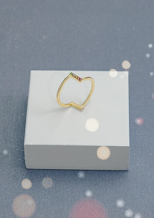 Anillo de oro de corazón abierto con pequeñas piedras preciosas multicolores en un lado, dispuestas sobre una plataforma cuadrada blanca sobre un fondo gris con efectos de luz. Esta impresionante pieza es parte de nuestra colección Joyas en Oro Laminado 18K.