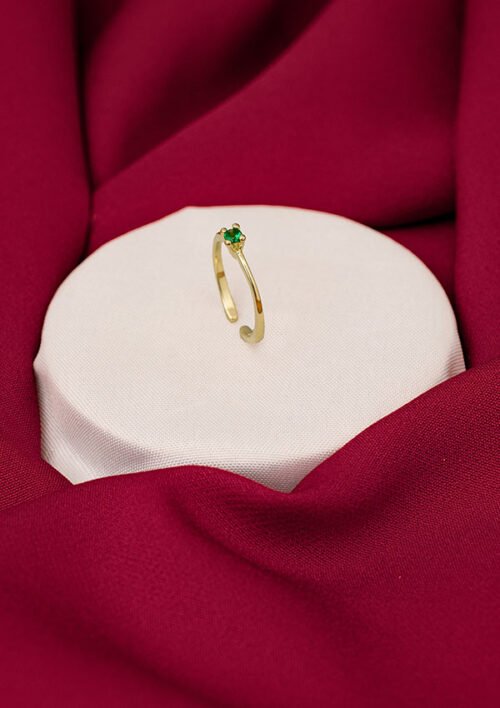 Un anillo de oro con una pequeña piedra preciosa verde se exhibe sobre una plataforma circular blanca, rodeada de tela roja. Esta exquisita pieza ejemplifica la elegancia de Joyas en oro laminado de 18k.