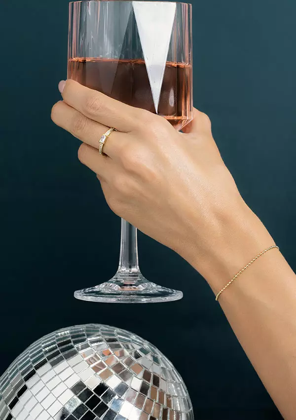 Una mano adornada con un anillo y pulsera de Joyas en Oro Laminado de 18K sostiene una copa de vino llena de líquido rosa. Una bola de discoteca con efecto espejo brilla en la parte inferior.