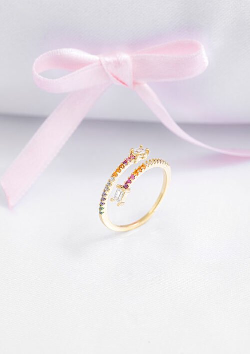 Un anillo de oro con un arco iris de pequeñas piedras preciosas y un lazo de cinta rosa en el fondo, presentado sobre una superficie blanca, muestra la elegancia de las joyas en oro laminado de 18k.