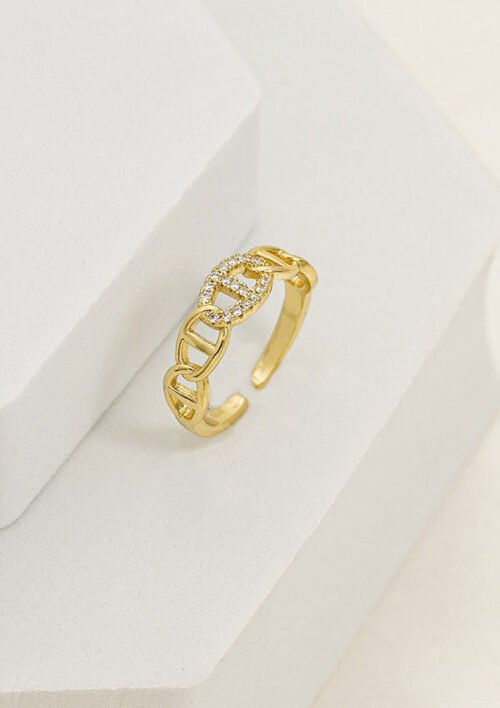 Un anillo de oro con diseño de eslabones de cadena y pequeños diamantes incrustados se muestra sobre una superficie geométrica blanca, mostrando la elegancia de las joyas en oro laminado de 18k.