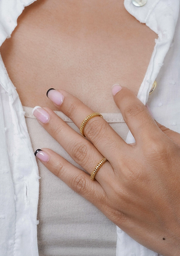 Una mano con uñas con puntas rosas y negras que lleva dos anillos de oro en los dedos índice y medio, mostrando impresionantes joyas en oro laminado de 18 quilates, colocadas sobre una parte superior beige y una tela texturizada blanca.