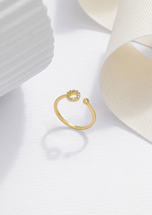 Un anillo de oro con un diseño de extremo abierto, que presenta dos pequeños grupos de diamantes, descansa sobre una superficie blanca texturizada junto a una cinta blanca. Esta exquisita pieza de joyería en oro laminado de 18k irradia elegancia y encanto atemporales.