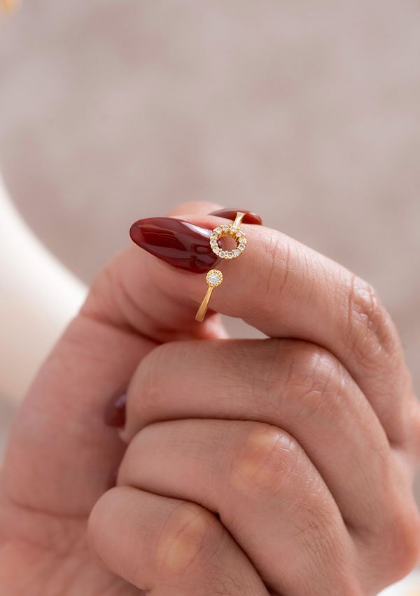 Una mano con uñas rojas cuidadas sosteniendo un anillo de oro que presenta un diseño circular con piedras preciosas incrustadas, mostrando la elegancia de las joyas en oro laminado de 18k.