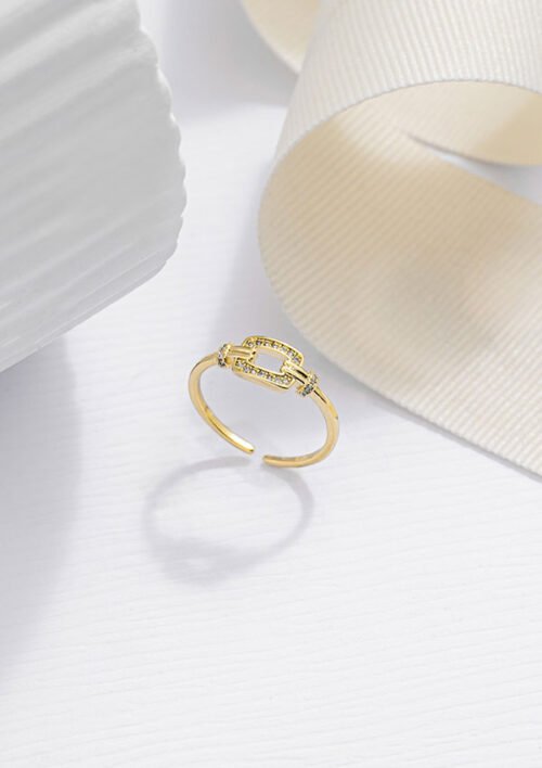 Un anillo de oro con diseño rectangular de rayos de sol reposa sobre una superficie blanca junto a una cinta de color crema, mostrando la elegancia de las joyas en oro laminado de 18k.