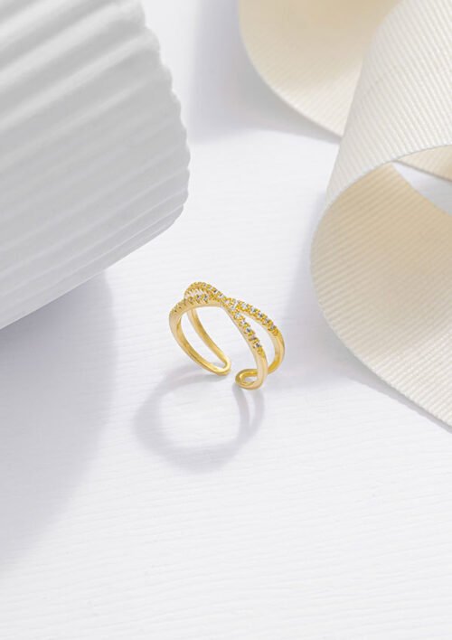 Un anillo de oro con diseño de doble banda y pequeños diamantes incrustados, elaborado con joyas en oro laminado de 18k, se muestra sobre un fondo blanco texturizado con cinta blanca.