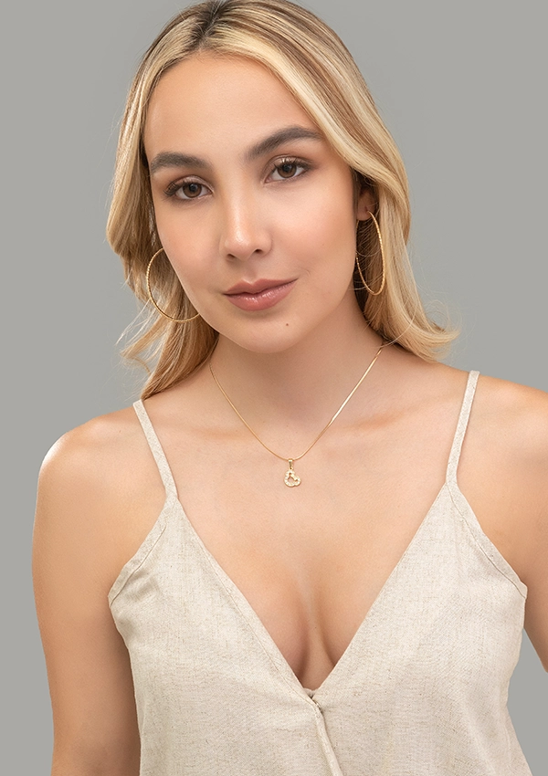 Una mujer de cabello largo y rubio que luce aretes de aro, un delicado collar con un colgante elaborado con joyas de oro laminado de 18 quilates y un top de cuello en V de color beige se encuentra sobre un fondo gris.