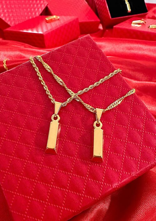 Dos collares colgantes de barra de oro con cadenas de cuerda se muestran en una caja acolchada roja con más cajas rojas en el fondo, mostrando la elegancia de las joyas en oro laminado de 18k.