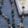 Un rosario con cuentas verdes y doradas se encuentra sobre una superficie texturizada. Presenta una cruz decorada con gemas verdes y una pequeña imagen dorada de una persona, capturando la elegancia de las joyas en oro laminado de 18k.