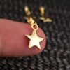 Primer plano de la punta del dedo de una persona que sostiene un pequeño dije dorado en forma de estrella sobre un fondo oscuro con textura. Dos dijes similares de "Joyas en Oro Laminado 18K" se ven borrosos en el fondo.