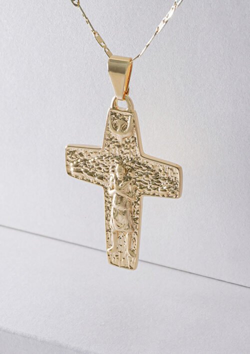 Colgante de cruz de oro con una figura grabada en su centro, colgando de una cadena de oro, bellamente elaborado como joyas en oro laminado de 18k, mostrado sobre un fondo claro.