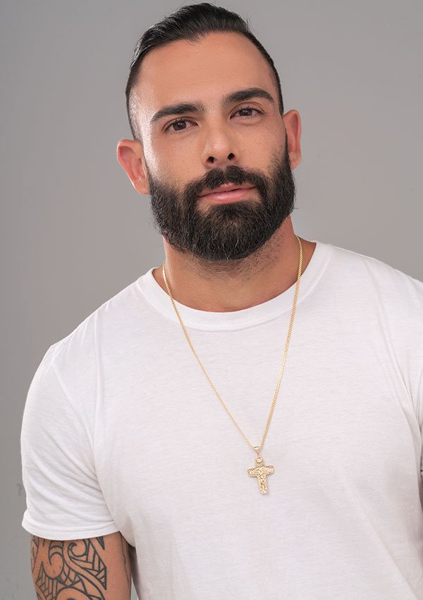 Un hombre con barba que viste una camisa blanca y un collar de oro con un colgante de cruz hecho con joyas en oro laminado de 18k. Tiene el pelo corto y un tatuaje en el brazo.