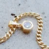 Primer plano de un collar de cadena de oro con un colgante hexagonal que presenta una única piedra preciosa transparente, colocada sobre una superficie gris claro. La pieza brilla con la elegancia de Joyas en Oro Laminado 18K, agregando un toque de sofisticación.