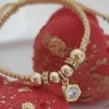 Una pulsera trenzada beige con cuentas de oro y un dije hexagonal que presenta un diamante, exhibido sobre piedras decorativas rojas y doradas, ejemplifica la elegancia de Joyas en Oro Laminado 18K.