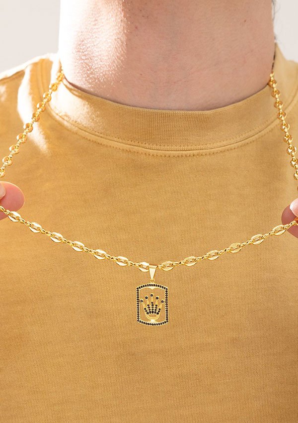 Una persona que viste una camisa amarilla sostiene un collar de cadena de oro con un colgante rectangular que presenta un emblema de corona, elaborado con joyas de oro laminado de 18k.