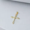 Un colgante de cruz de oro adornado con pequeñas piedras preciosas, que encarna la elegancia de Joyas en Oro Laminado 18K, colocado sobre una superficie texturizada blanca.