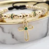 Una pulsera de cuentas de oro con detalles de piedras preciosas verdes, que presenta un dije de cruz, descansa sobre una superficie de mármol junto a otra pieza de la colección Joyas en Oro Laminado de 18K.