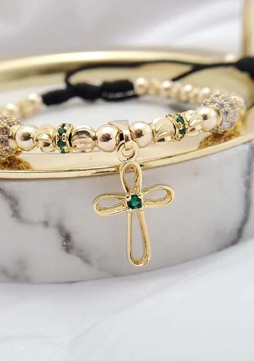 Una pulsera de cuentas de oro con detalles de piedras preciosas verdes, que presenta un dije de cruz, descansa sobre una superficie de mármol junto a otra pieza de la colección Joyas en Oro Laminado de 18K.