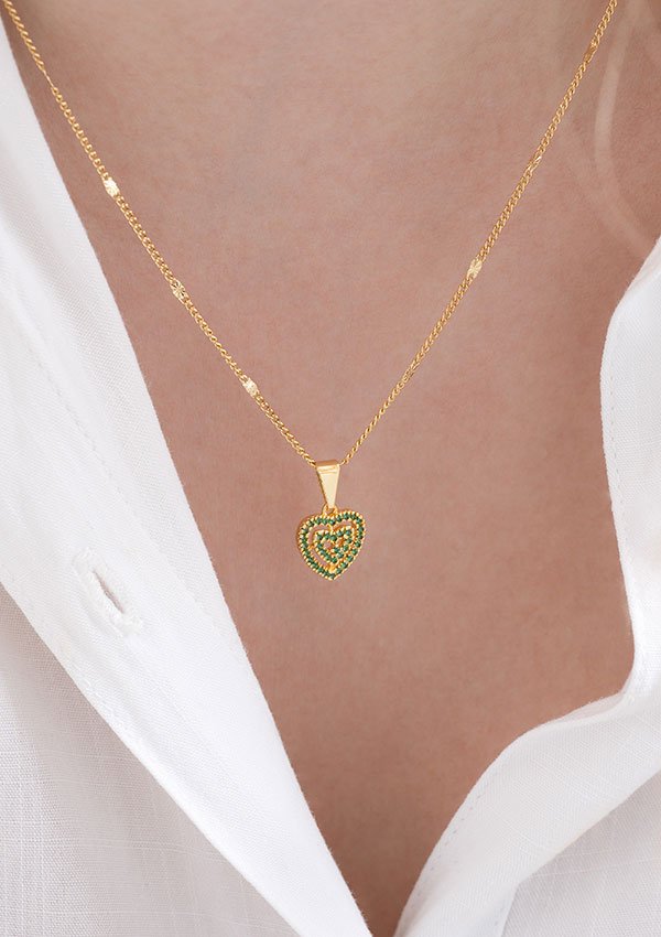 Una persona que lleva un collar de oro con un colgante en forma de corazón adornado con piedras preciosas verdes, resaltado sobre una camisa blanca. Esta exquisita pieza es parte de nuestra colección Joyas en oro laminado de 18k.