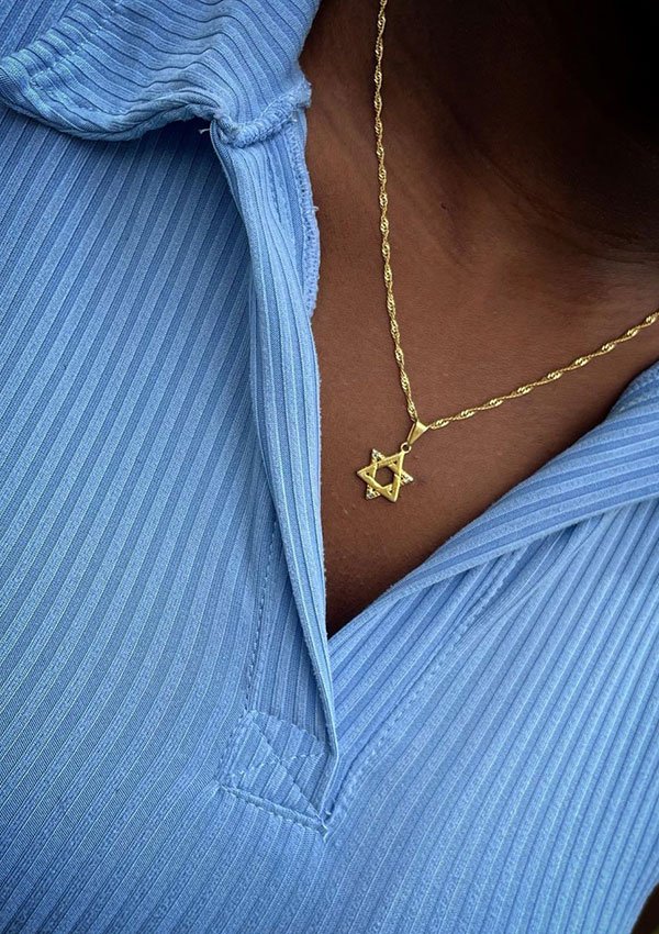 Primer plano de una persona que viste una camisa de cuello acanalado de color azul claro con un collar de estrella de David de oro elaborado con joyas en oro laminado de 18k.