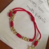 Una pulsera de hilo rojo con cuentas de oro y un dije de estrella de David, elaborada con joyas en oro laminado de 18k, reposa sobre un papel blanco con texto escrito a mano en español.