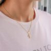 Una persona lleva un collar con un colgante de cruz elaborado con joyas de oro laminado de 18k y una camisa rosa claro con un texto parcial que dice "Nacido para ser".