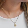 Una persona que lleva un collar de cadena de oro con un pequeño colgante ovalado que tiene una figura grabada elaborada con joyas de oro laminado de 18 quilates. La persona está vestida con una camiseta blanca.