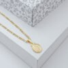 Un collar de cadena de oro con un colgante ovalado reposa sobre una superficie blanca, mostrando la elegancia de las joyas en oro laminado de 18k, con una caja decorativa estampada en gris y blanco en el fondo.