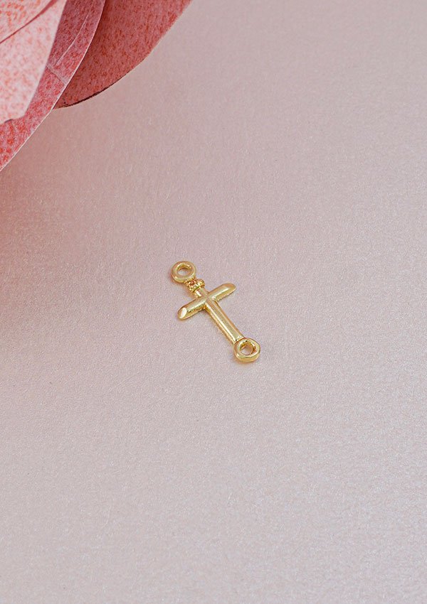 Un colgante Herraje Cruz Inri 1.5 Cms, una pequeña cruz de oro con dos presillas elaborada en oro laminado de 18k (joyas en oro laminado 18k), se coloca sobre una superficie beige suave cerca de un material texturizado rosa.