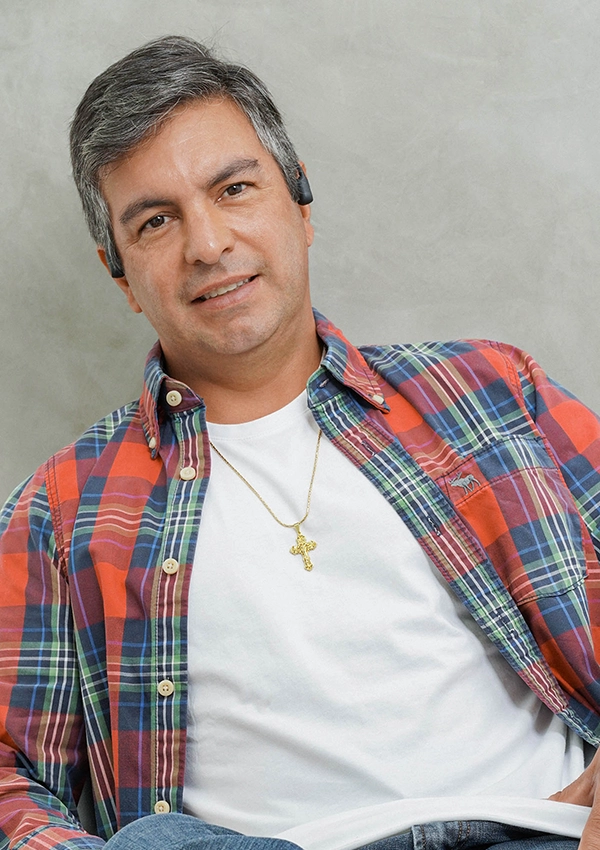 Hombre de mediana edad, de cabello corto y gris, que viste una camisa a cuadros roja sobre una camiseta blanca, un collar de oro con una cruz de la colección Joyas en Oro Laminado 18K y un auricular, sonriendo mientras está sentado sobre un fondo neutro.