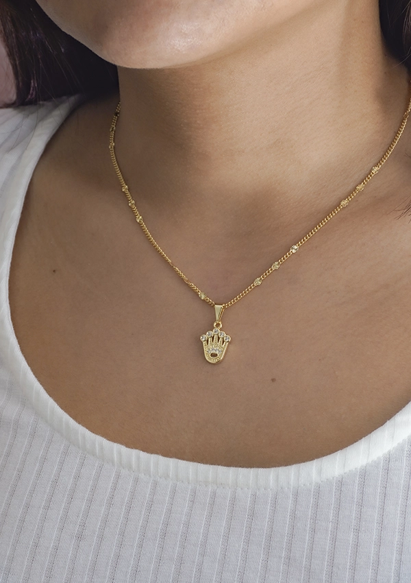 Primer plano de una persona que lleva un collar de oro con un colgante en forma de mano. La persona está vestida con una camisa blanca de canalé, mostrando la elegancia de Joyas en Oro Laminado 18K.