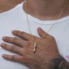 Primer plano de una persona que viste una camisa blanca y un collar de oro con un colgante que se asemeja a una mancuerna, elaborado con joyas de oro laminado de 18k. La persona tiene un tatuaje en el antebrazo y su mano descansa sobre su pecho.