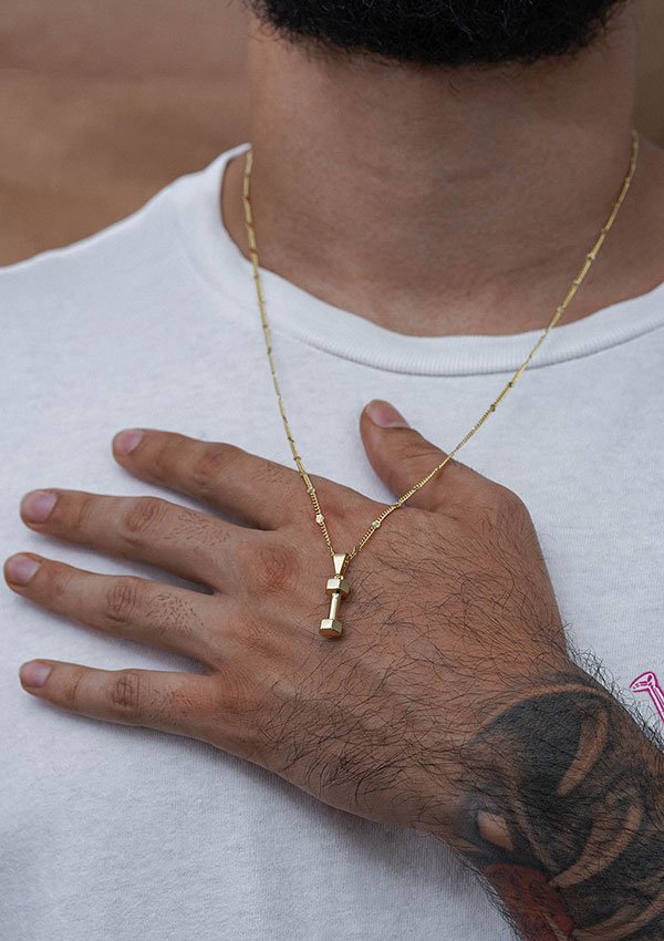 Primer plano de una persona que viste una camisa blanca y un collar de oro con un colgante que se asemeja a una mancuerna, elaborado con joyas de oro laminado de 18k. La persona tiene un tatuaje en el antebrazo y su mano descansa sobre su pecho.