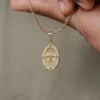 Un primer plano de la mano de una persona sosteniendo un collar de oro con un colgante ovalado de diseño intrincado, que muestra la exquisita artesanía de Joyas en Oro Laminado 18K.