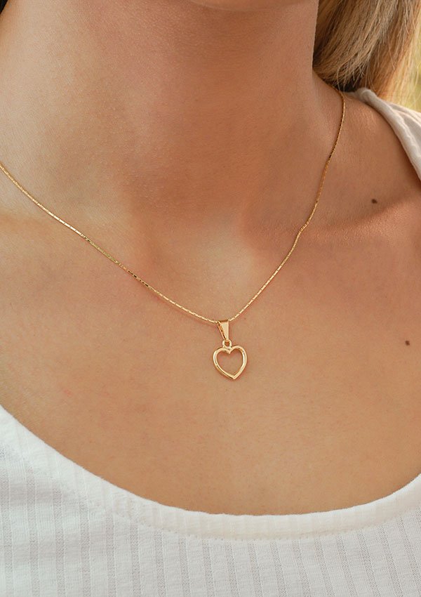 Una persona que lleva un fino collar de oro con el Dije Corazón Silueta, elaborado con joyas en oro laminado de 18k, sobre una parte superior acanalada de color claro.