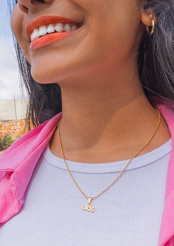 Una mujer que lleva un collar de oro con las letras "Dije Dios", con joyas en oro laminado de 18k, una camisa blanca y una chaqueta rosa sonríe al aire libre.