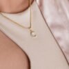 Primer plano de una persona que lleva el *Dije Media Luna Circonias*, un collar colgante de media luna de oro elaborado con joyas en oro laminado de 18k, combinado con un top beige claro con escote de cerradura y una chaqueta de satén de color claro.