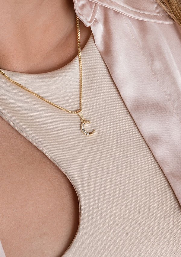 Primer plano de una persona que lleva el *Dije Media Luna Circonias*, un collar colgante de media luna de oro elaborado con joyas en oro laminado de 18k, combinado con un top beige claro con escote de cerradura y una chaqueta de satén de color claro.