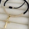 El Dije Cruz de la Vida, una pulsera de cordón ajustable negro adornada con pequeñas cuentas de oro y un colgante ankh dorado elaborado con joyas bañadas en oro de 18k, se exhibe sobre una superficie acolchada.