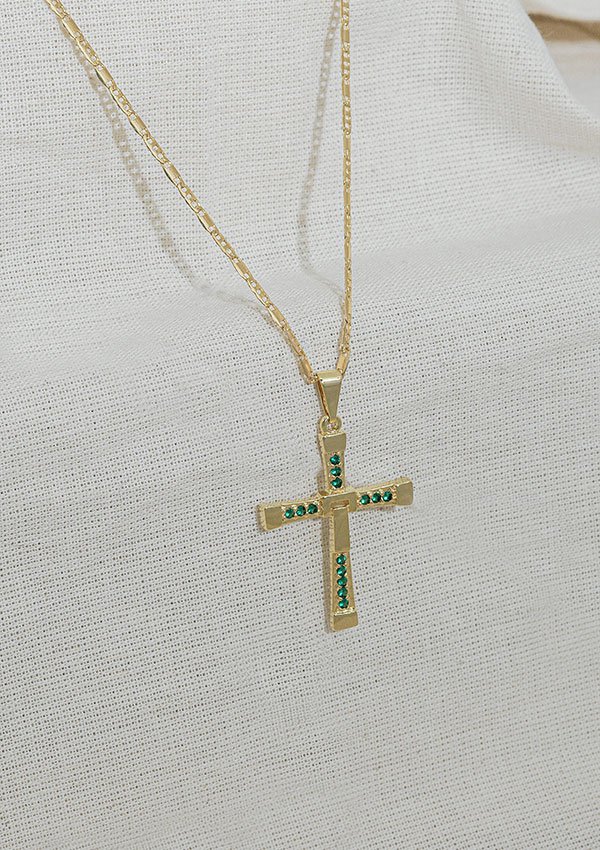 Un colgante de cruz de oro, Dije Cruz Toretto 3,1 Cms, con piedras verdes en una cadena de oro exhibida sobre un fondo de tela muestra la elegancia de las joyas en oro laminado de 18k.