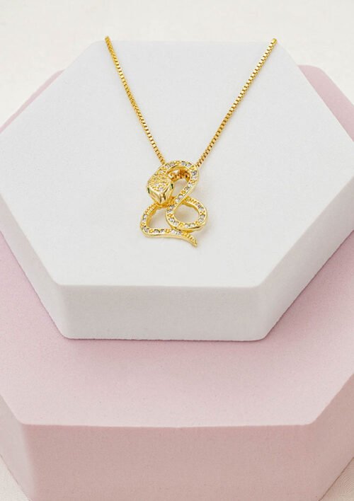 Dije Snake, un collar de oro que presenta un colgante en forma de serpiente adornado con pequeños diamantes, elegantemente expuesto sobre un joyero hexagonal blanco con base rosa. Perfecto para quienes aprecian la elegancia de las joyas en oro laminado de 18k.