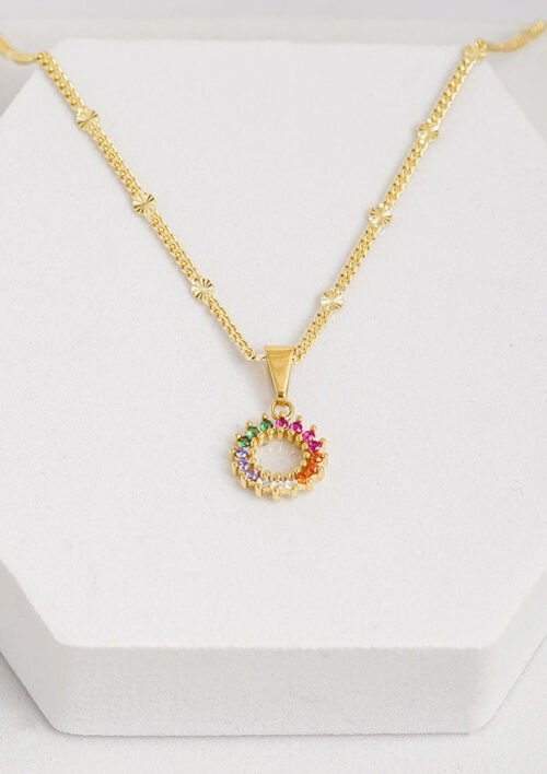 Sobre un fondo blanco se exhibe un collar de oro con cadena de pequeñas formas de estrellas y un colgante circular adornado con piedras preciosas multicolores. Es una de las exquisitas joyas en oro laminado de 18k que irradia elegancia y encanto.