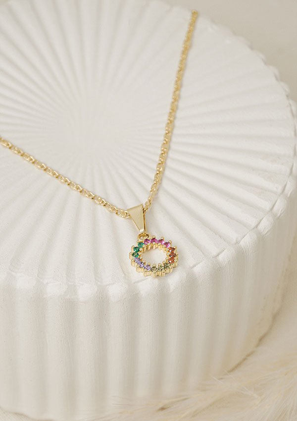 Collar de oro con colgante que presenta un pequeño diseño circular adornado con piedras preciosas multicolores. La pieza, realizada con joyas en oro laminado de 18k, se exhibe sobre una superficie estriada de color blanco.