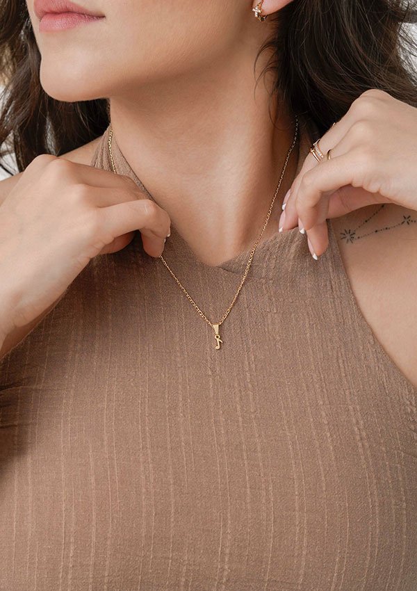 Una persona que viste una blusa beige sin mangas acanalada toca un pequeño collar de oro con un colgante. El cabello oscuro de la persona enmarca su rostro y sus orejas tienen piercings adornados con pequeños aretes, mostrando elegantes joyas en oro laminado de 18k.