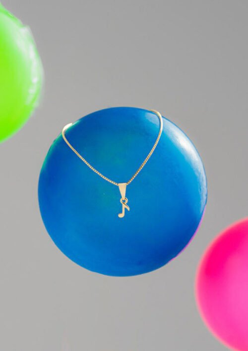 Un collar de oro con un colgante de nota musical se encuentra sobre un globo azul flotante, con globos verdes y rosados parcialmente visibles en el fondo. El collar, parte de nuestra colección Joyas en oro laminado de 18k, agrega un toque elegante a esta exhibición extravagante.