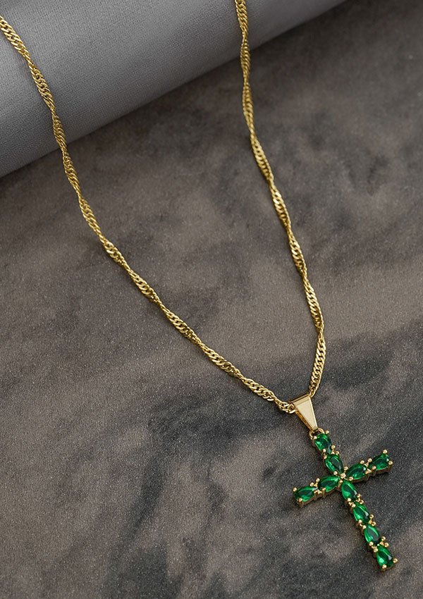 Collar de oro con colgante de cruz adornado con piedras preciosas verdes vibrantes, exhibidas sobre un fondo con textura de piedra, ejemplificando la elegancia de las joyas en oro laminado de 18k.