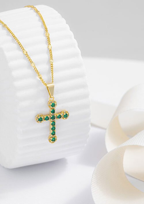 Colgante de cruz de oro con incrustaciones de piedras preciosas verdes en una cadena de oro, que se muestra sobre un fondo cilíndrico blanco con una cinta blanca cerca. Esta exquisita pieza es parte de nuestra colección Joyas en oro laminado de 18k, que ofrece elegancia y sofisticación.