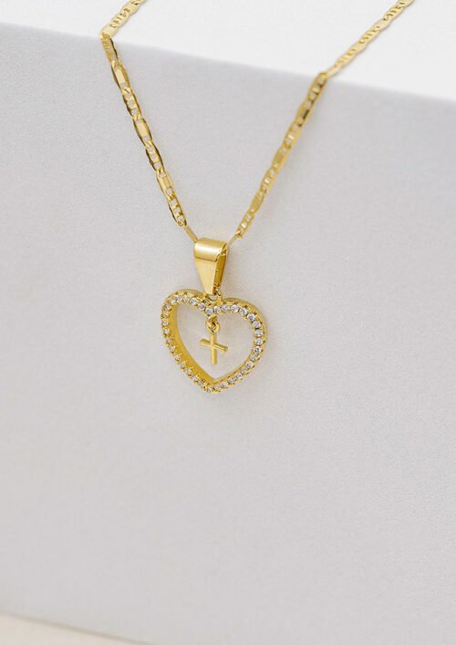 Un collar de oro con un colgante en forma de corazón que presenta una cruz, adornada con pequeños cristales, colocados sobre una superficie gris claro: joyas en oro laminado de 18k que destilan elegancia y encanto.